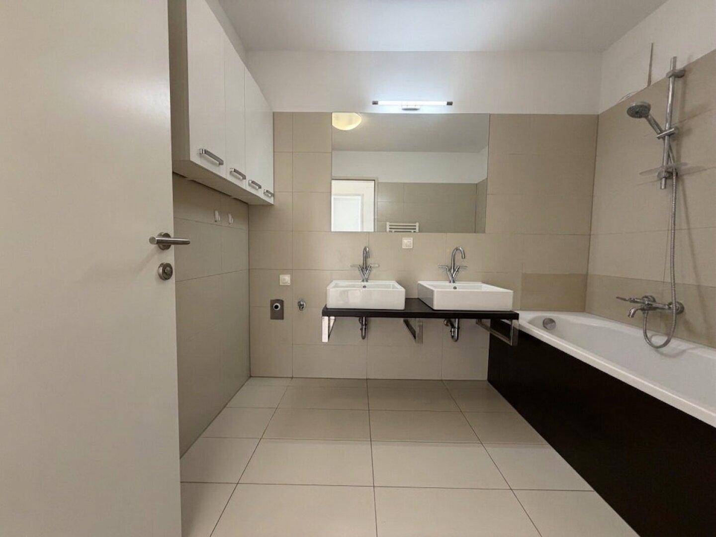 Modernes Badezimmer mit Doppelwaschbecken, Badewanne und hellen Fliesen an Wänden und Boden.