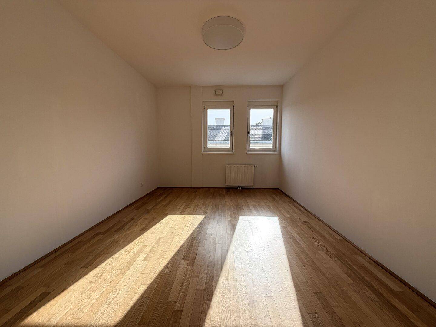 Helles, leeres Zimmer mit Holzboden und zwei Fenstern, die viel Tageslicht hereinlassen.