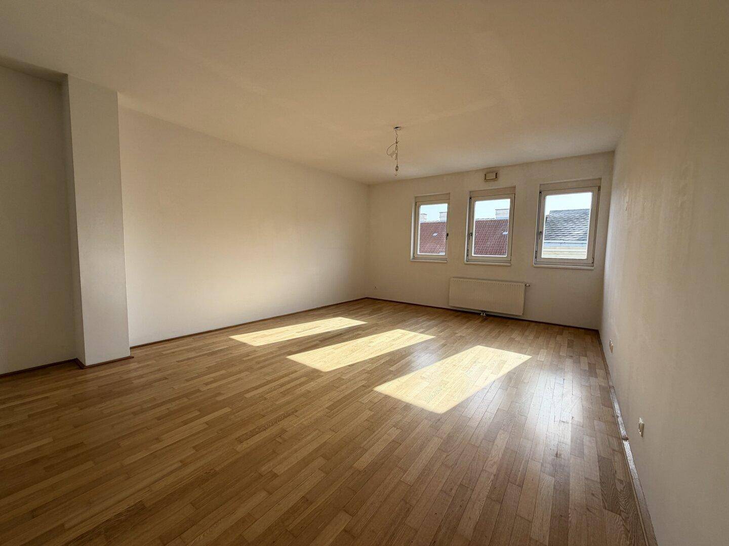 Geräumiges, leeres Zimmer mit Holzboden und viel natürlichem Licht durch drei Fenster.