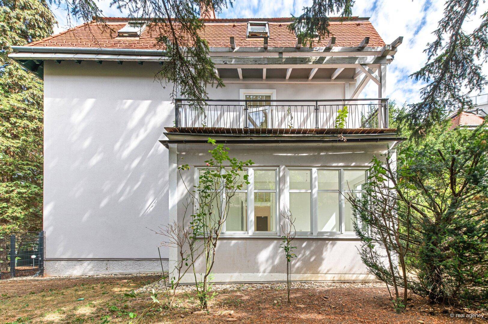 Rückansicht des Gebäudes mit Balkon und Gartenbereich, umgeben von Bäumen.