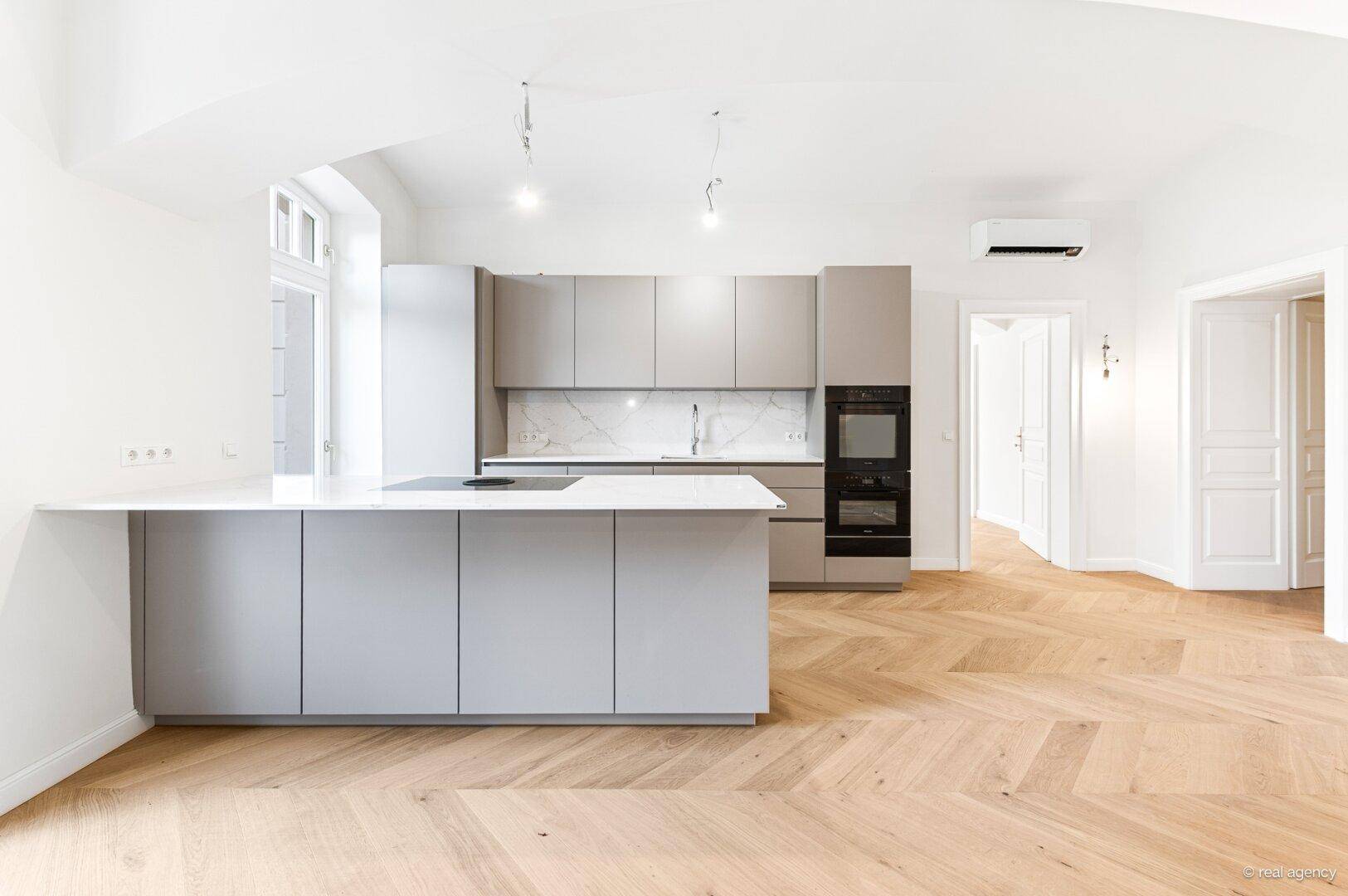 Moderne Einbauküche mit Kücheninsel, grauen Fronten und hochwertigen Geräten in offenem Wohnraum.