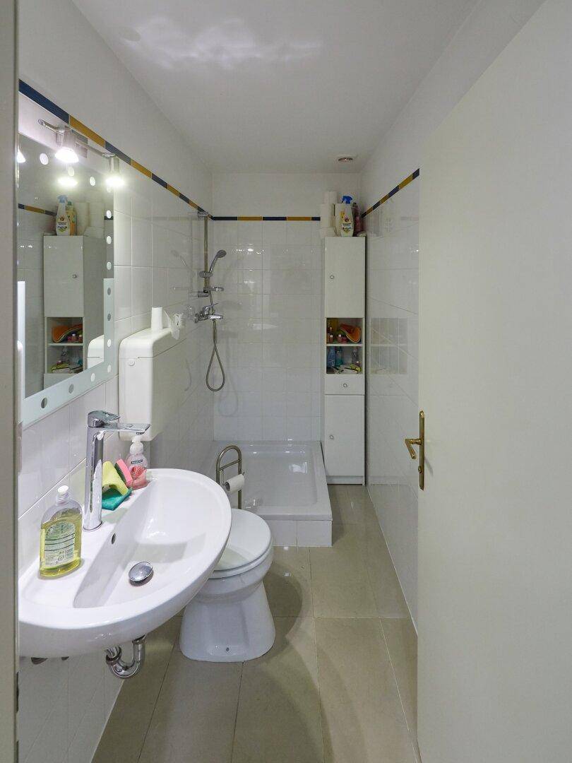 Schlichtes Badezimmer mit Waschbecken, Toilette und Duschbereich, ausgestattet mit weißen Fliesen.