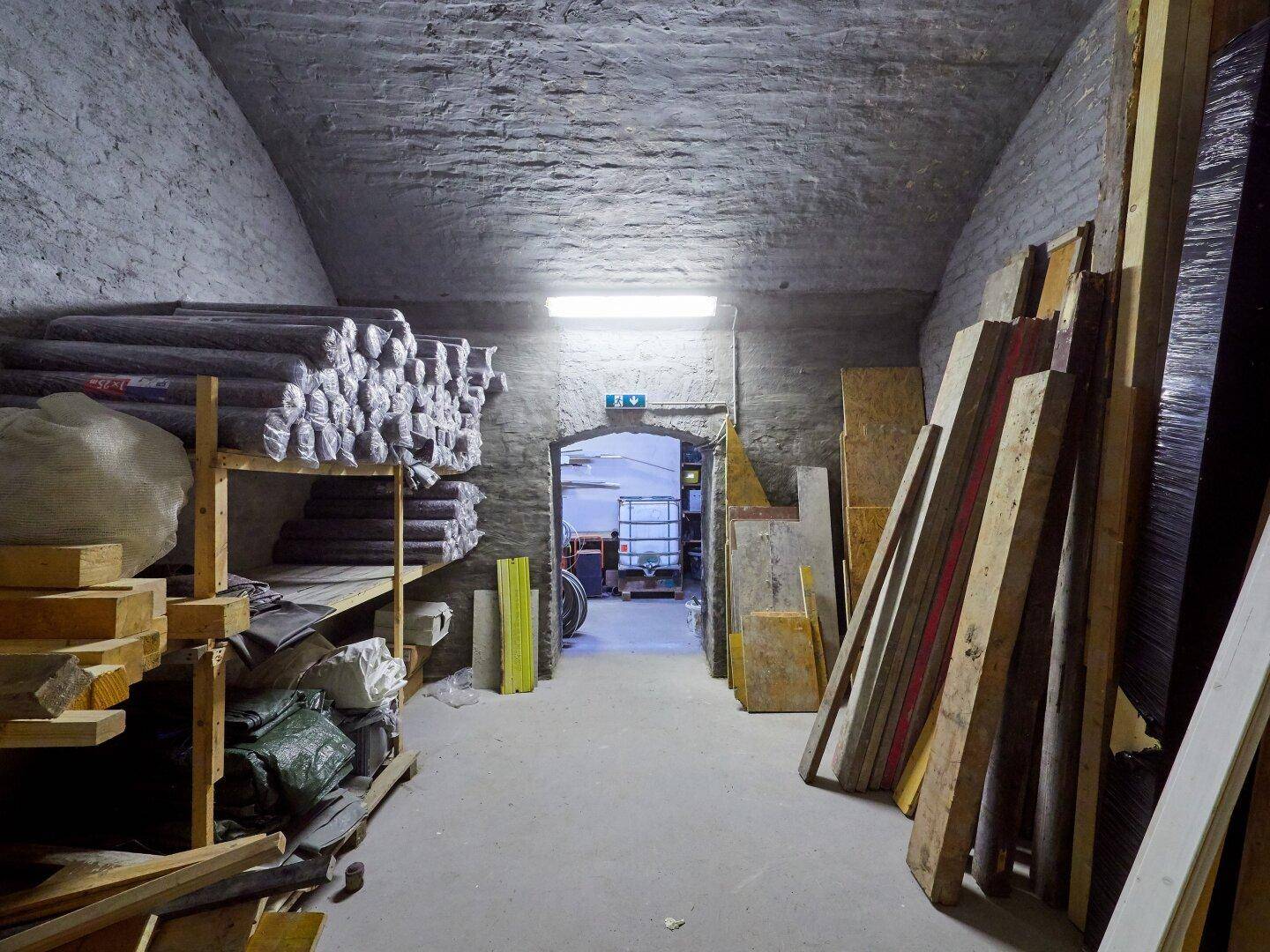 Langer, schmaler Kellergang mit gelagertem Holz und Baumaterialien, beleuchtet von Neonröhren.