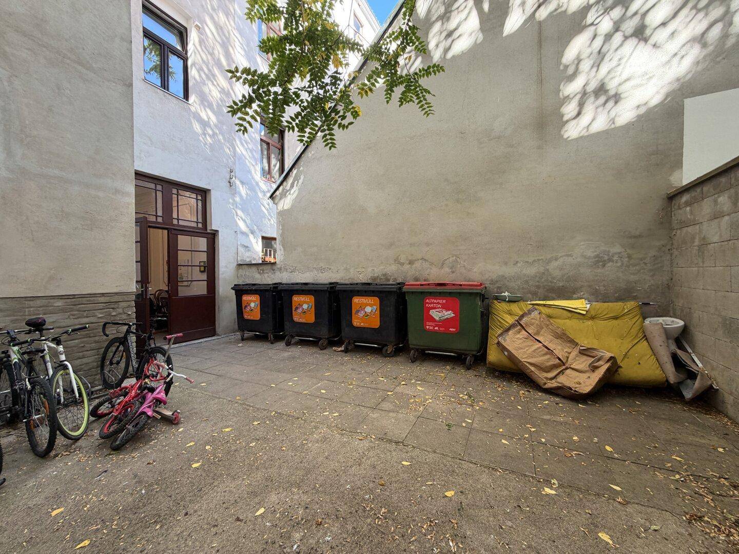 Außenbereich mit Mülltonnen, Fahrrädern und einer Gebäudefassade im Hintergrund.