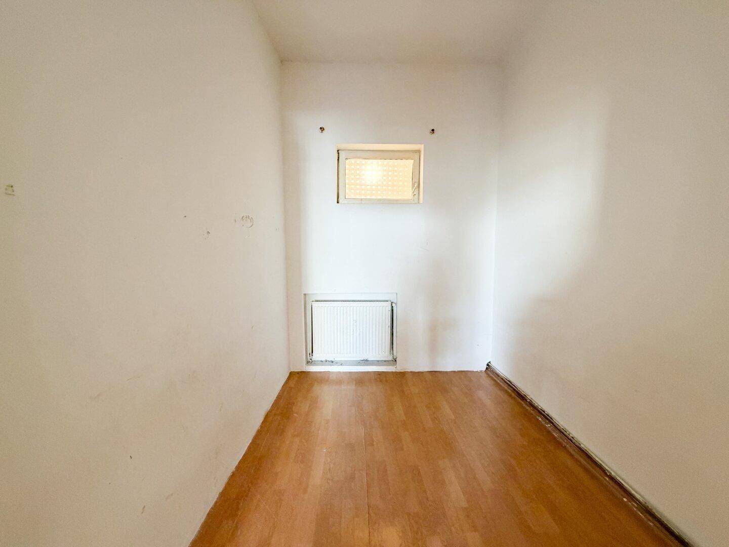 Kleiner Raum mit Holzboden, einem hochgelegenen Fenster und Heizkörper.