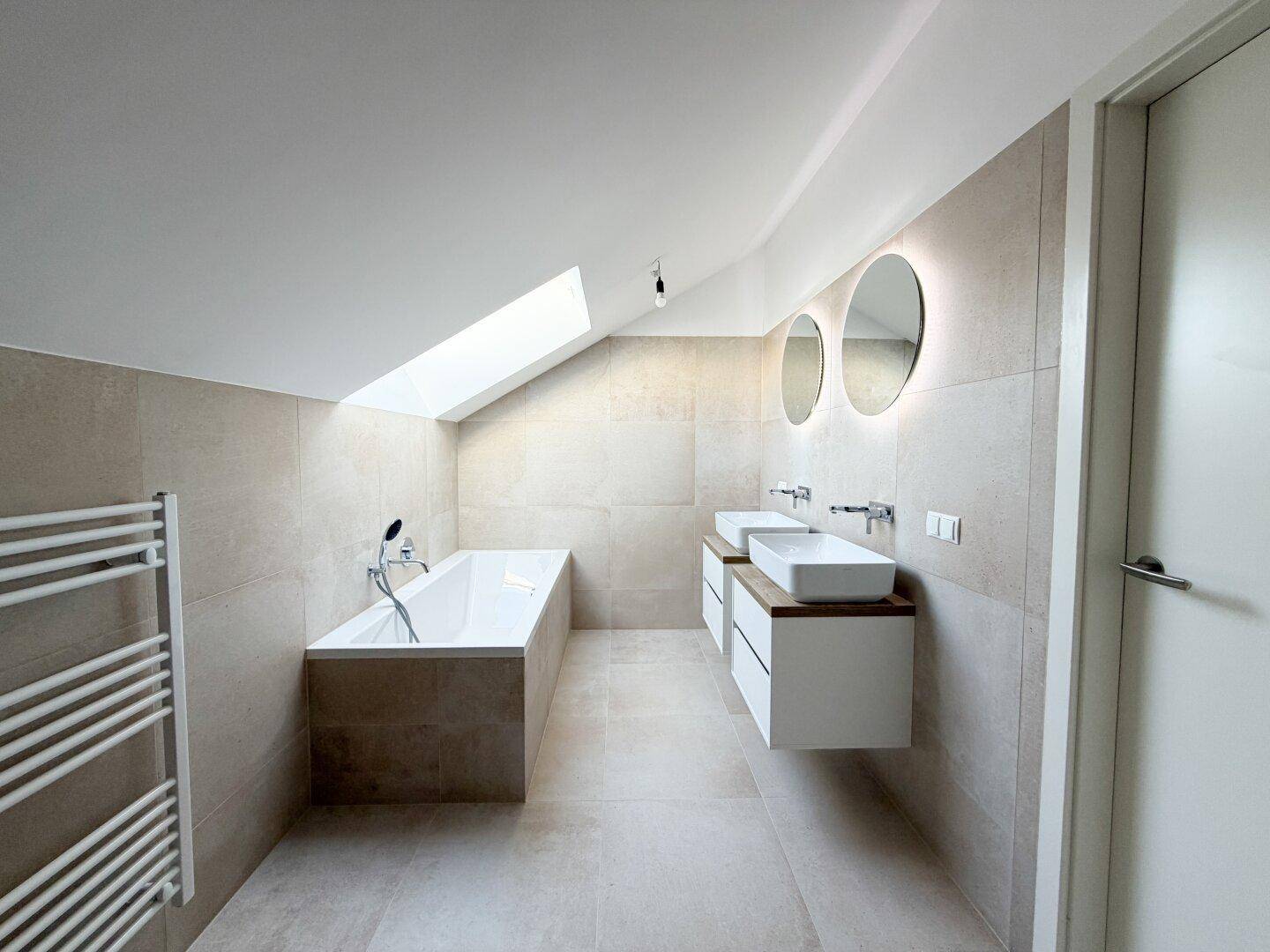 Helles Badezimmer mit beigefarbenen Fliesen, Doppelwaschbecken, Badewanne und Dachfenster.