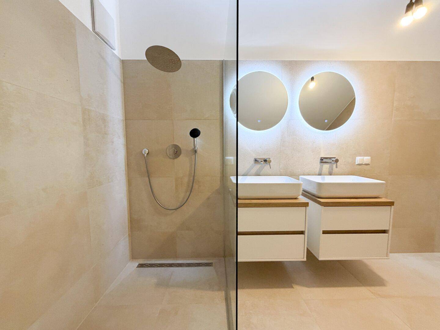 Begehbare Dusche mit Regenduschkopf und Doppelwaschbecken mit beleuchteten Spiegeln.
