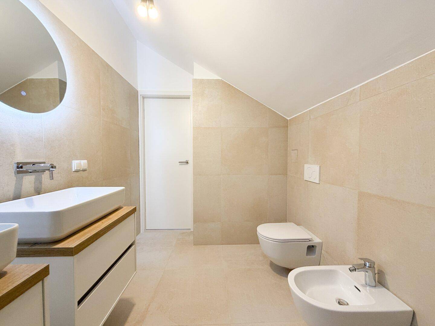 Modernes Badezimmer mit Doppelwaschbecken, Wand-WC, Bidet und beigefarbenen Fliesen.