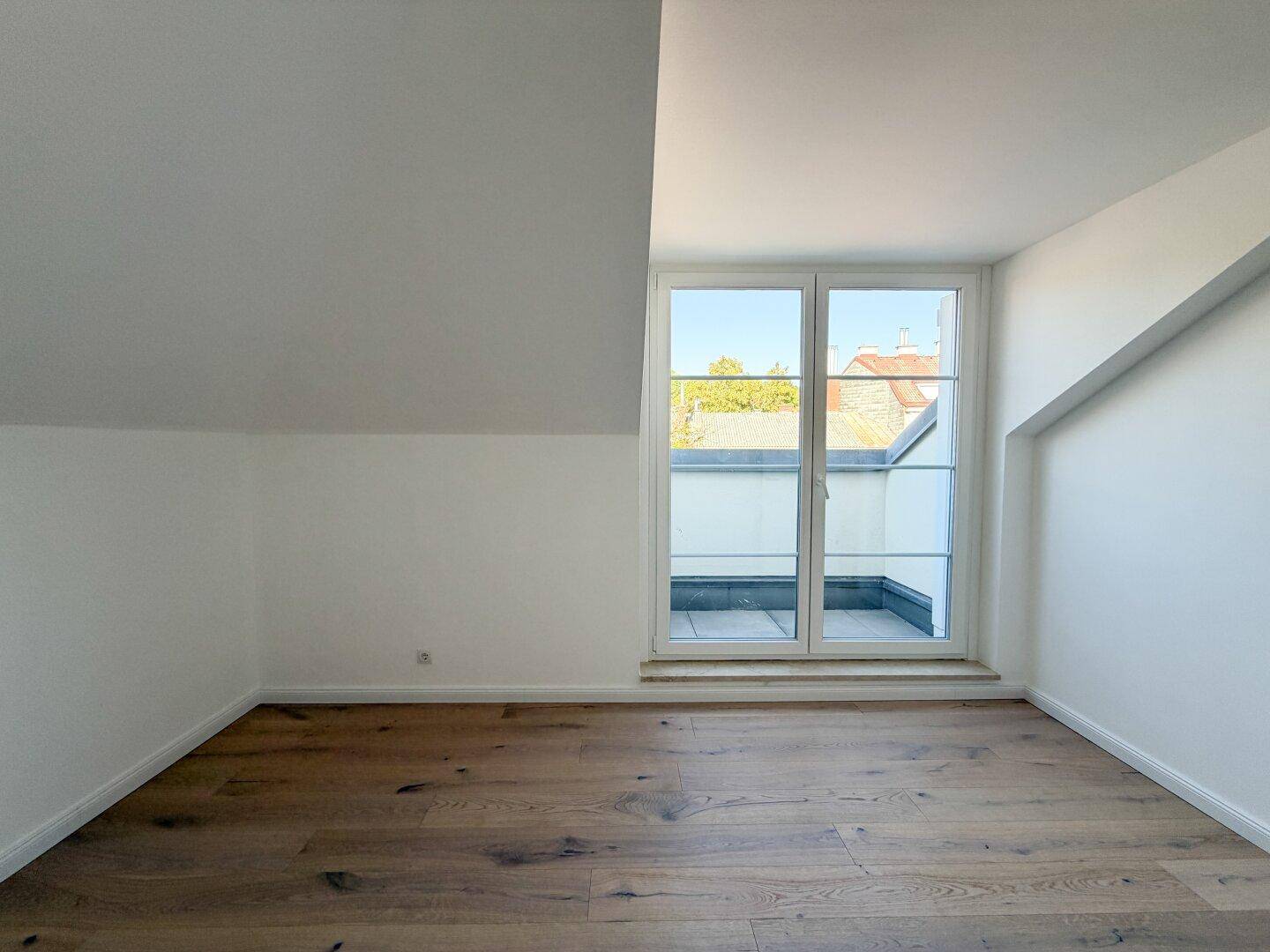 Helles Zimmer mit Holzboden, schrägen Decken und Zugang zu einem kleinen Balkon.
