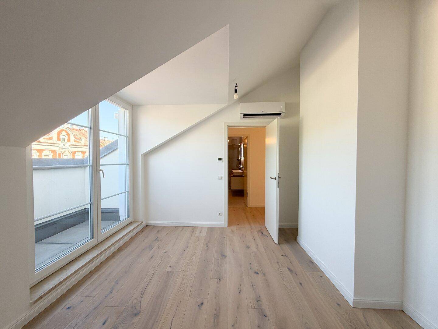 Helles Schlafzimmer mit Holzboden, schrägen Decken und Zugang zum Balkon und Bad.