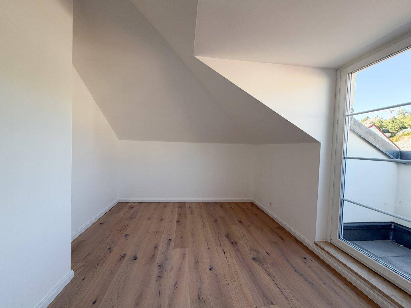 Geräumiges Zimmer mit Holzboden, schrägen Decken und Blick auf den kleinen Balkon.