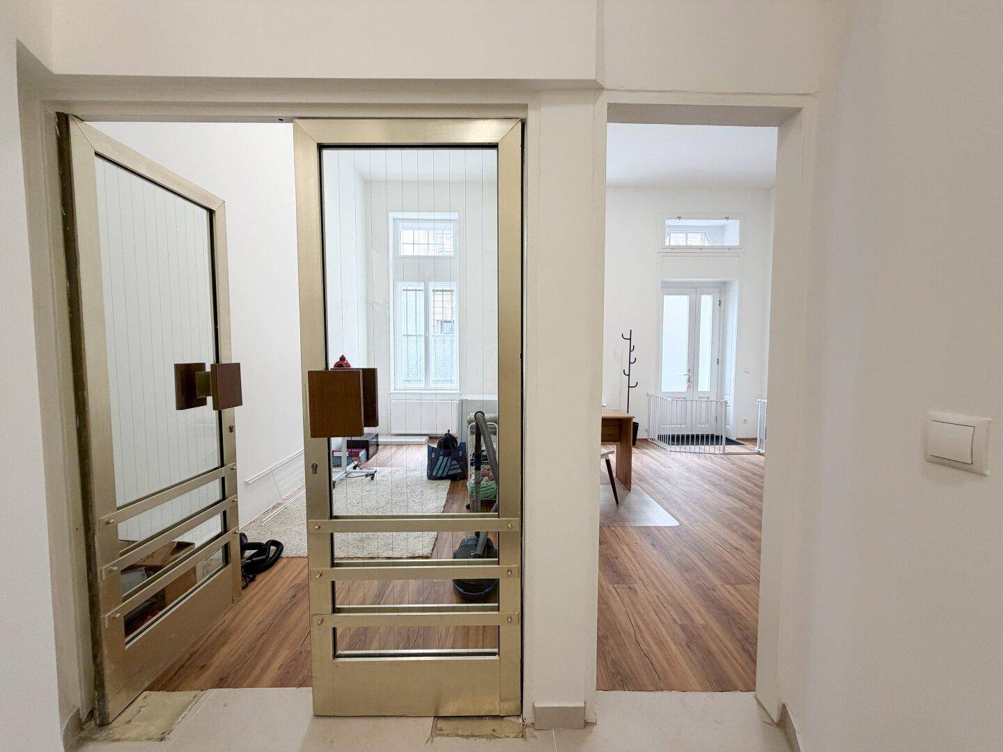 Blick durch eine goldfarbene Glastür in einen möblierten Geschäftsraum mit Holzboden und hohen Fenstern.