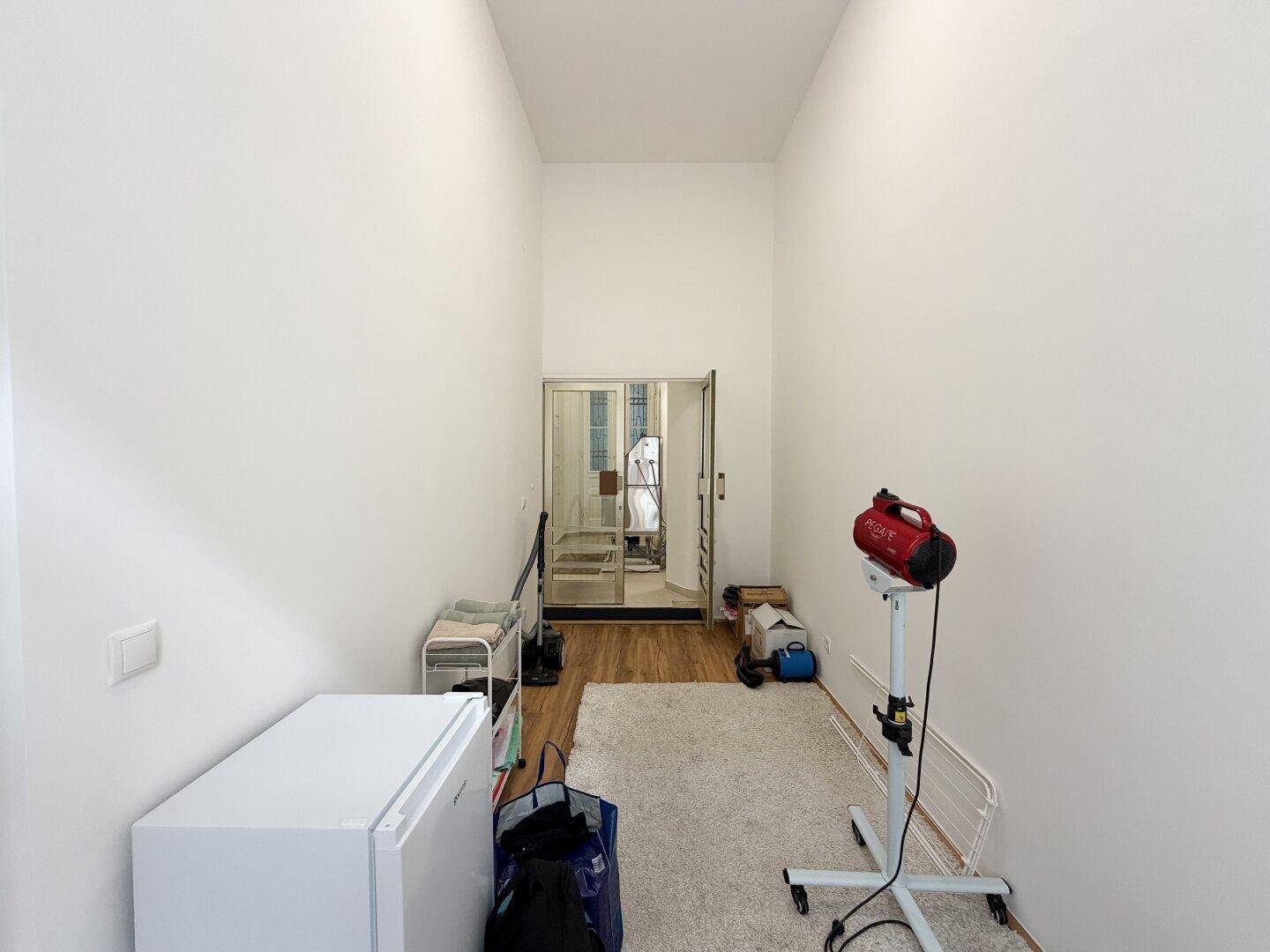 Langer, schmaler Raum mit Teppich, Kühlschrank und Arbeitsgeräten, mit Blick auf eine Glastür.