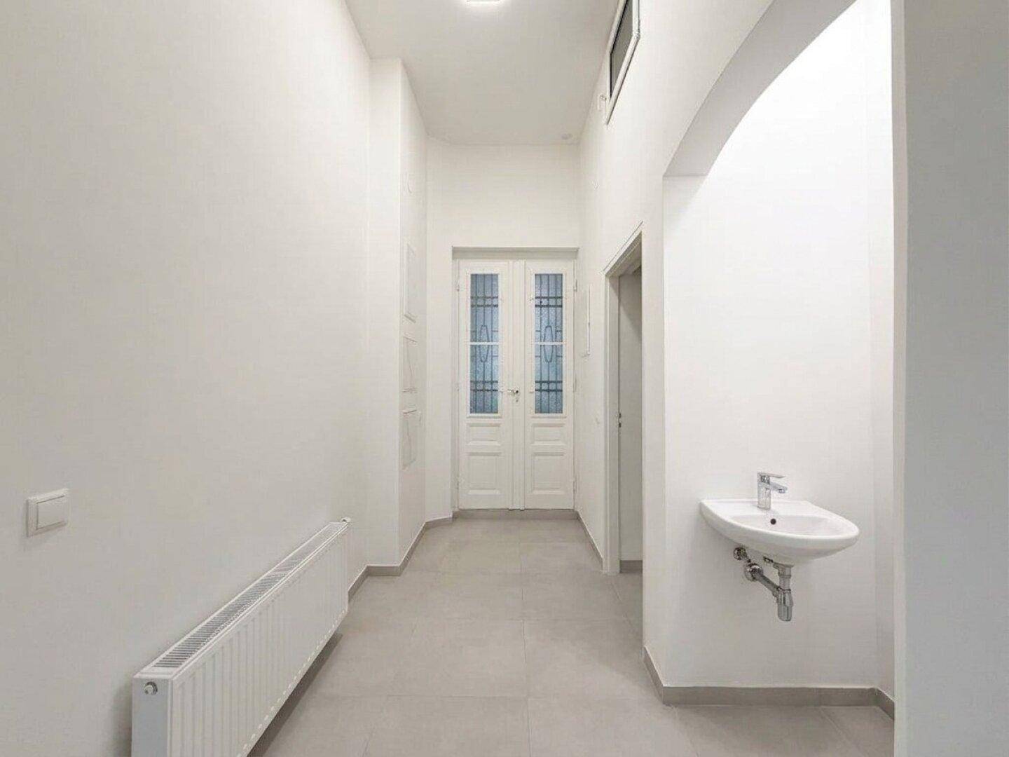 Heller Flur mit Fliesenboden, Heizkörper und einem kleinen Waschbecken an der Wand.