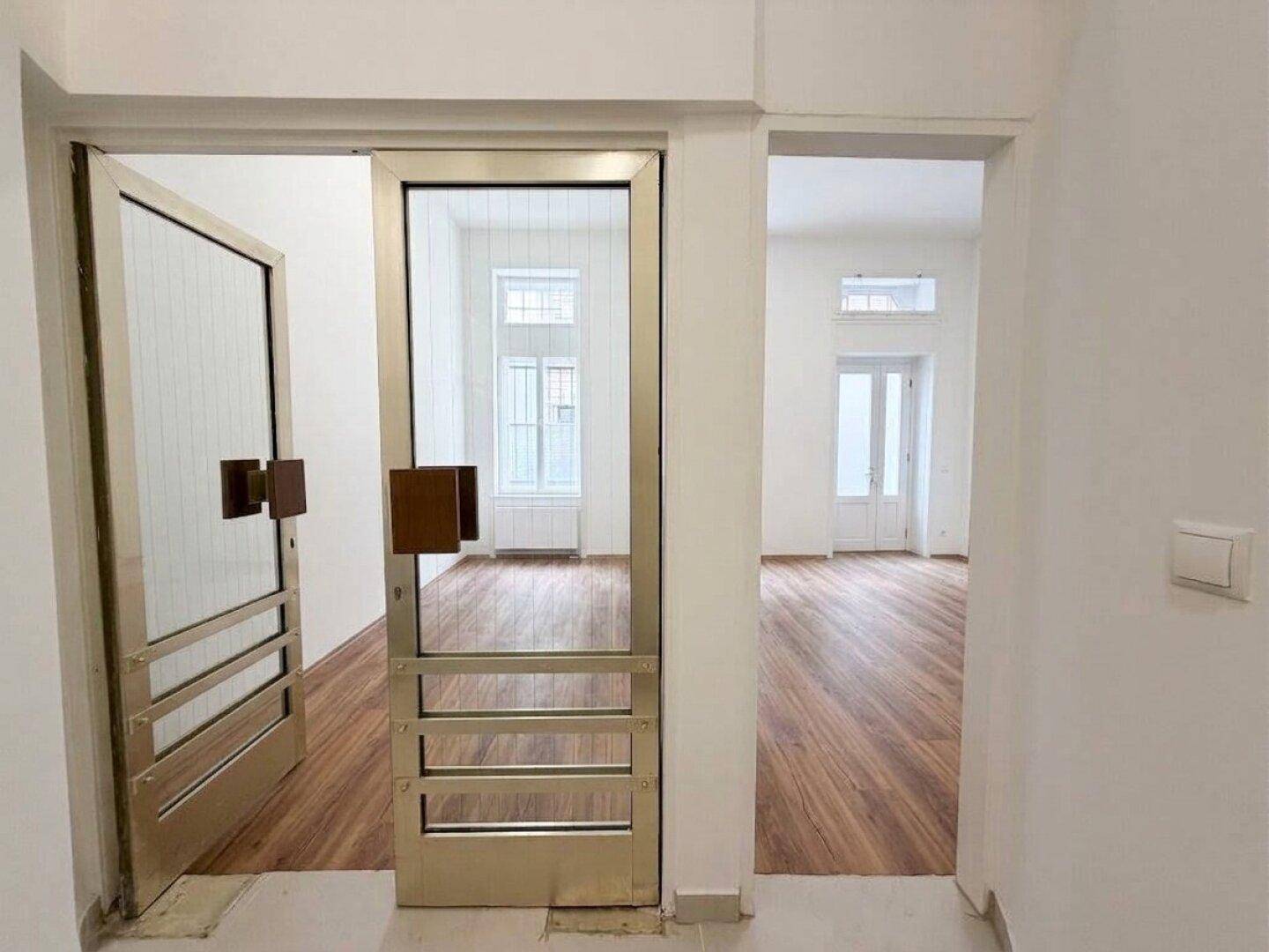 Blick durch eine goldfarbene Glastür in einen weiteren hellen Raum mit Holzboden und hohen Fenstern.