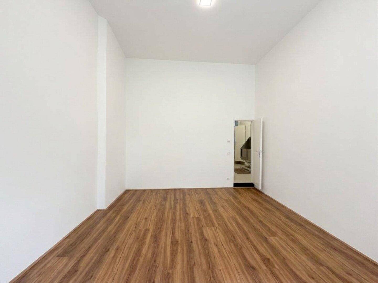 Langer, schmaler Raum mit hellem Laminatboden und weiß gestrichenen Wänden, mit Zugang zu einem weiteren Bereich.