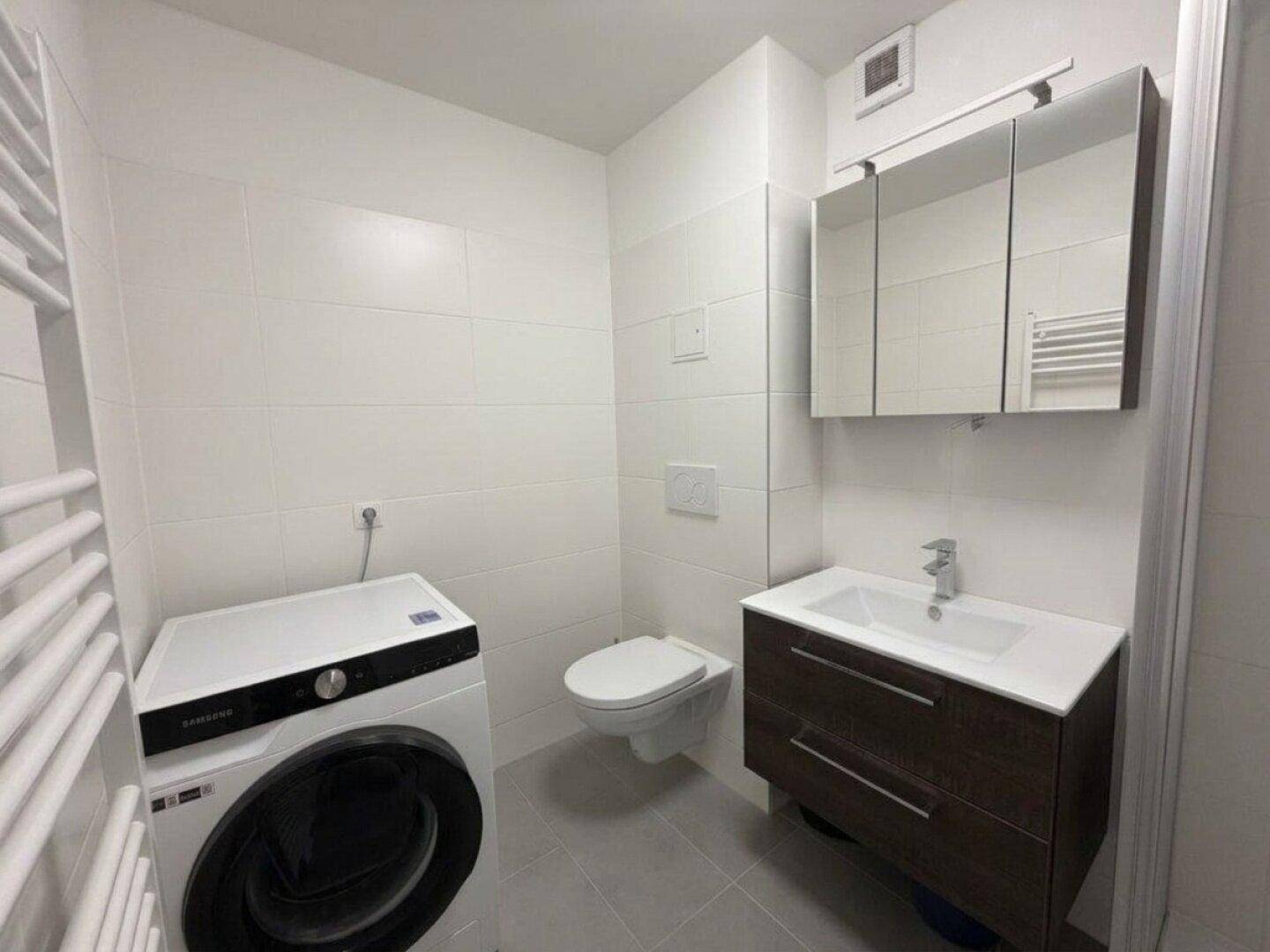 Modernes Badezimmer mit weißen Fliesen, Waschbecken, Toilette und Waschmaschine.