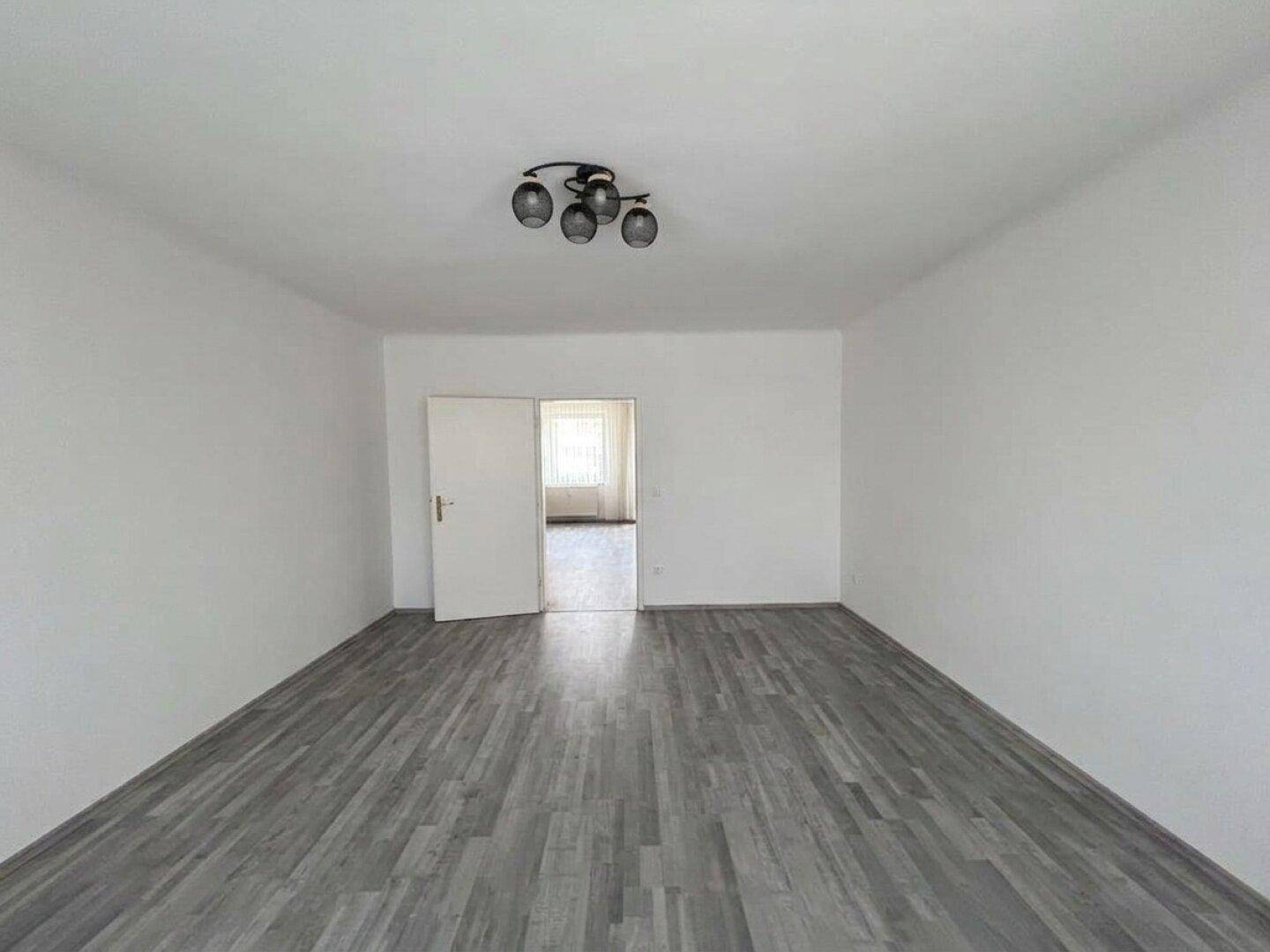 Leerer Wohnraum mit grauem Laminatboden und offener Tür zu einem weiteren Zimmer.