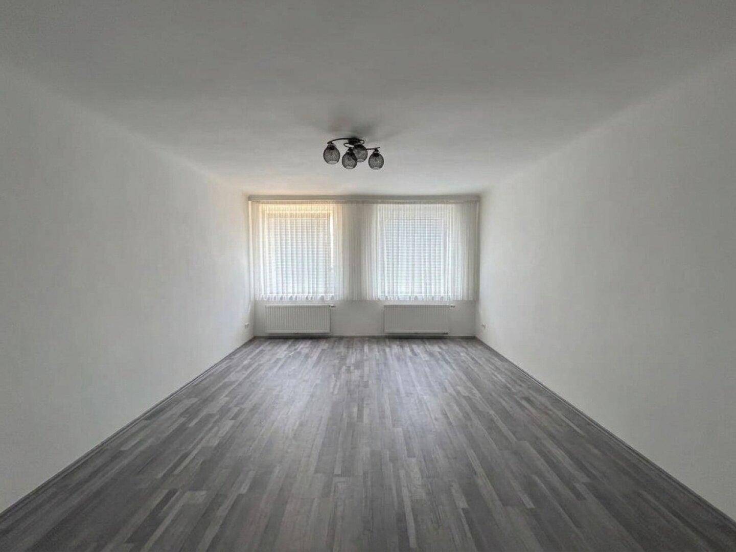 Geräumiger, heller Raum mit grauem Laminatboden und zwei großen Fenstern.