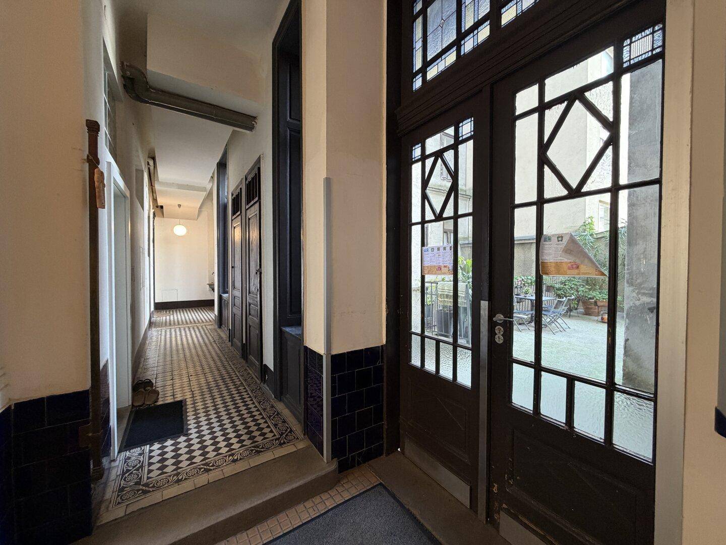 Historischer Flur mit gemusterten Fliesen und Blick durch verglaste Türen in einen Innenhof.