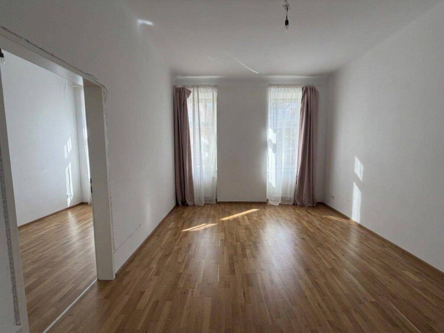 Helles, leeres Wohnzimmer mit Holzboden und zwei großen Fenstern, bereit zur Einrichtung.