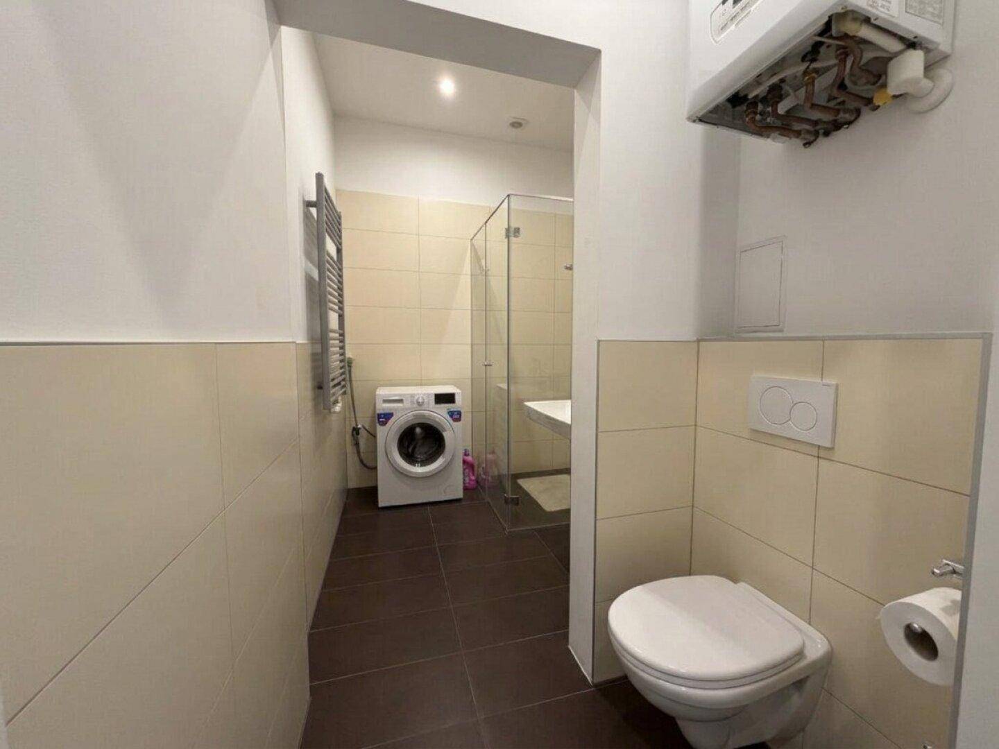 Badezimmer mit wandhängendem WC, Waschmaschine, Dusche und beheiztem Handtuchhalter.
