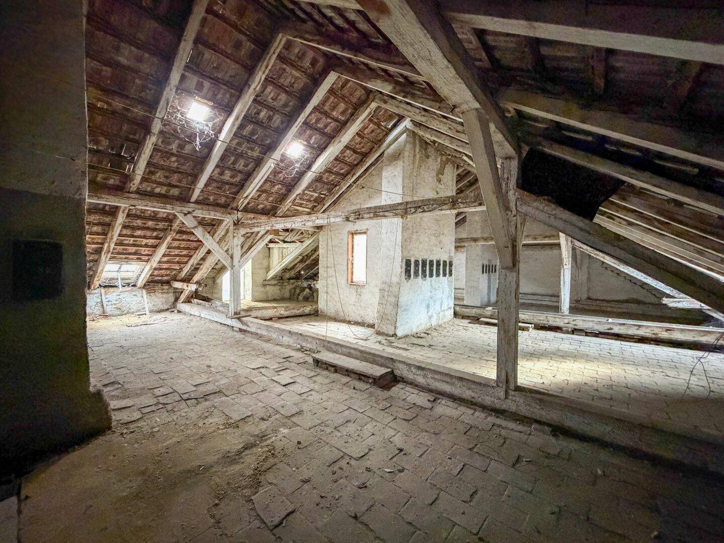 Weiter Blick in den ungenutzten Dachboden mit mehreren Dachfenstern, Holzbalken und Ziegelboden.
