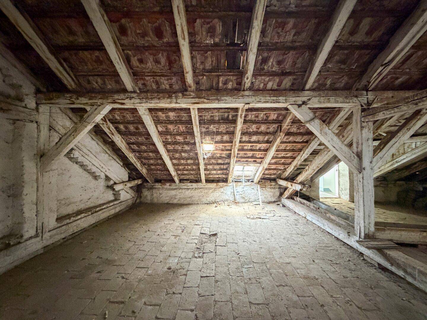 Geräumiger, unausgebauter Dachboden mit sichtbaren Holzbalken, Ziegelboden und einem Dachfenster.