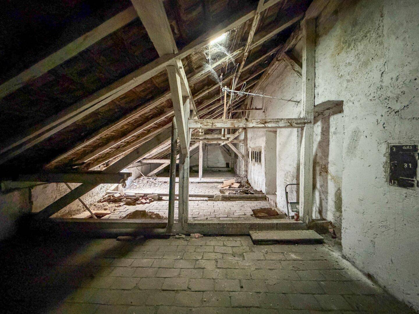 Unausgebauter Dachbodenbereich mit freiliegenden Holzbalken, Ziegelboden und rohen Wänden.