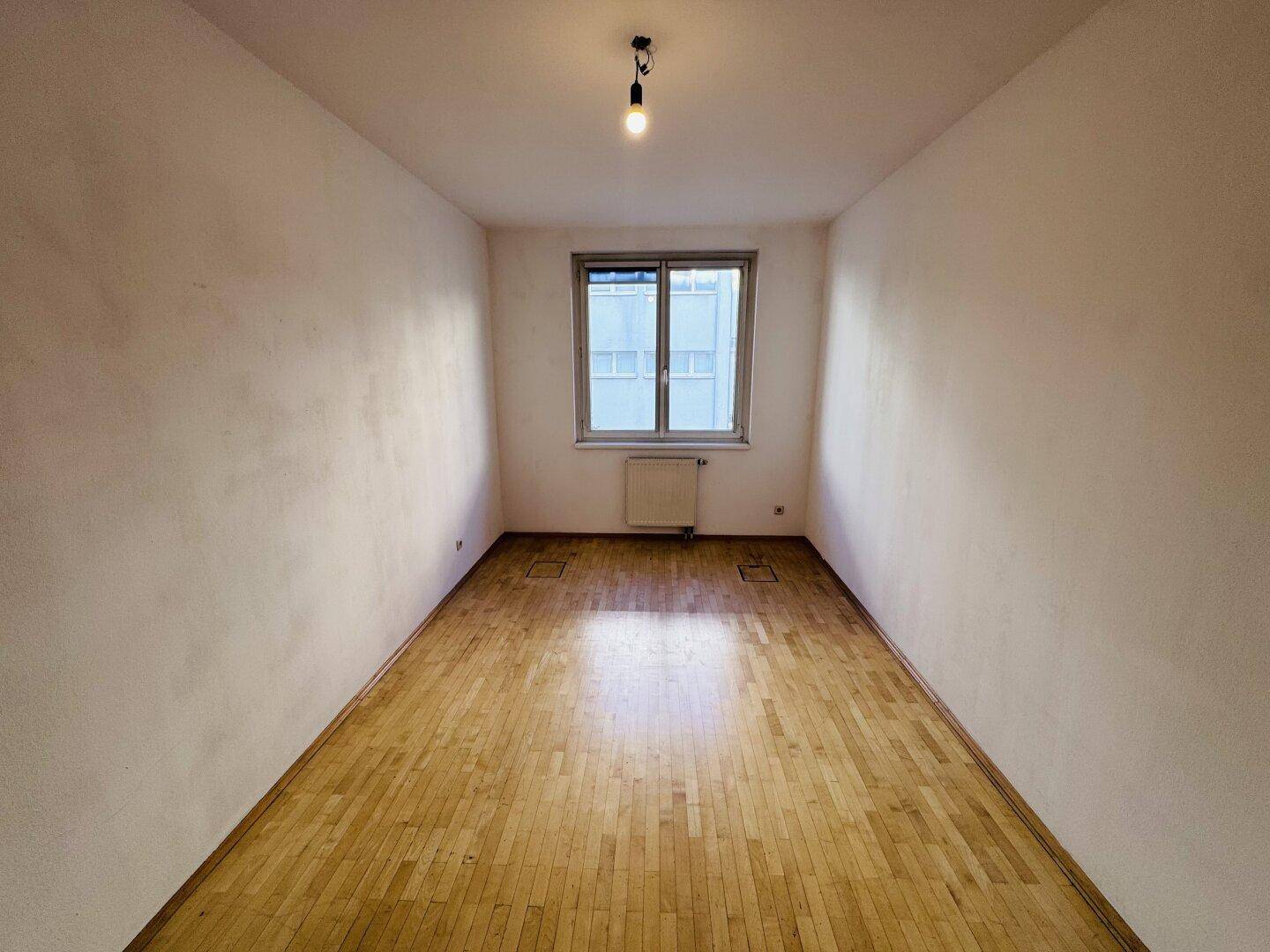 Heller, leerer Raum mit Holzboden und einem Fenster, das für natürliche Belichtung sorgt.