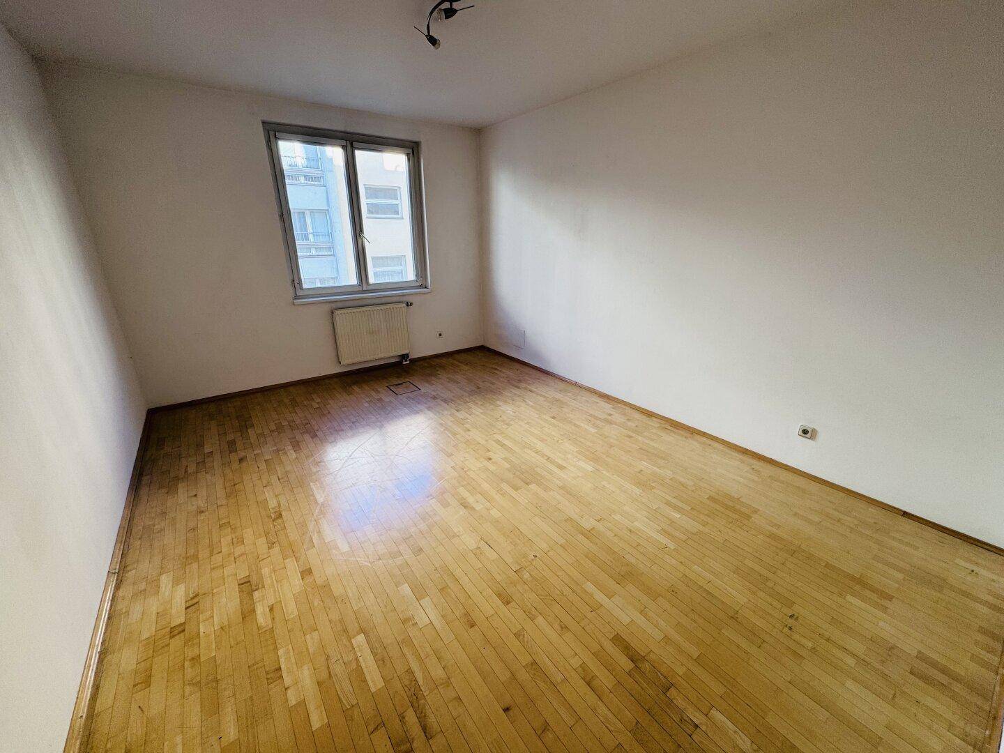 Geräumiger, leerer Raum mit Holzboden und einem großen Fenster, das viel Licht spendet.