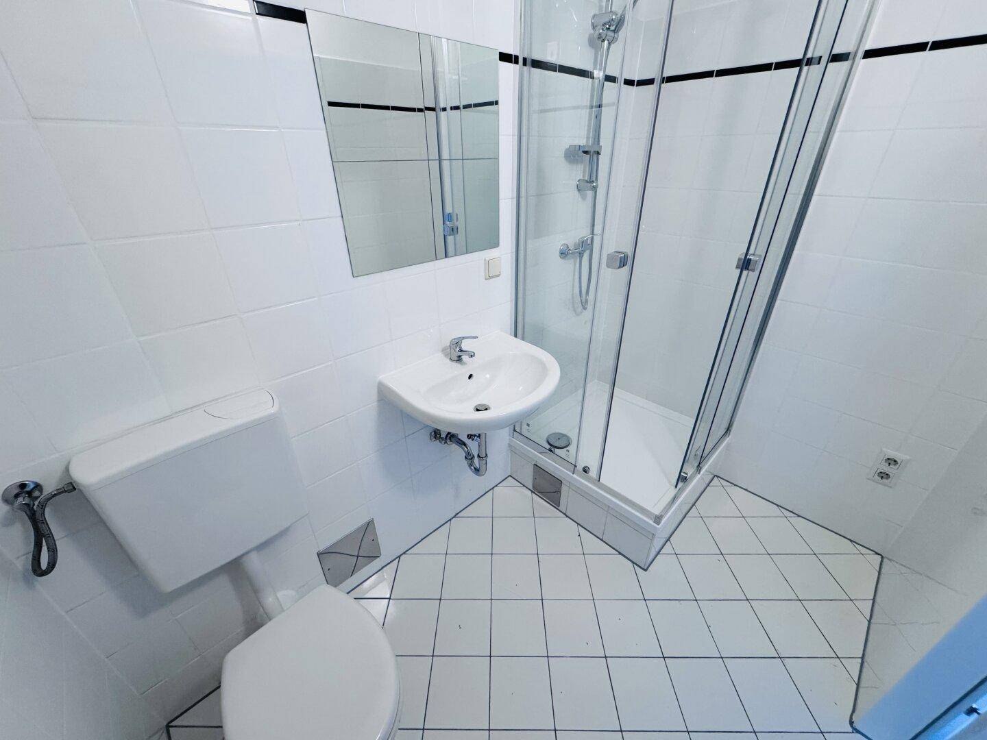 Kleines Badezimmer mit weißen Fliesen, Toilette, Eckwaschbecken und Duschkabine.