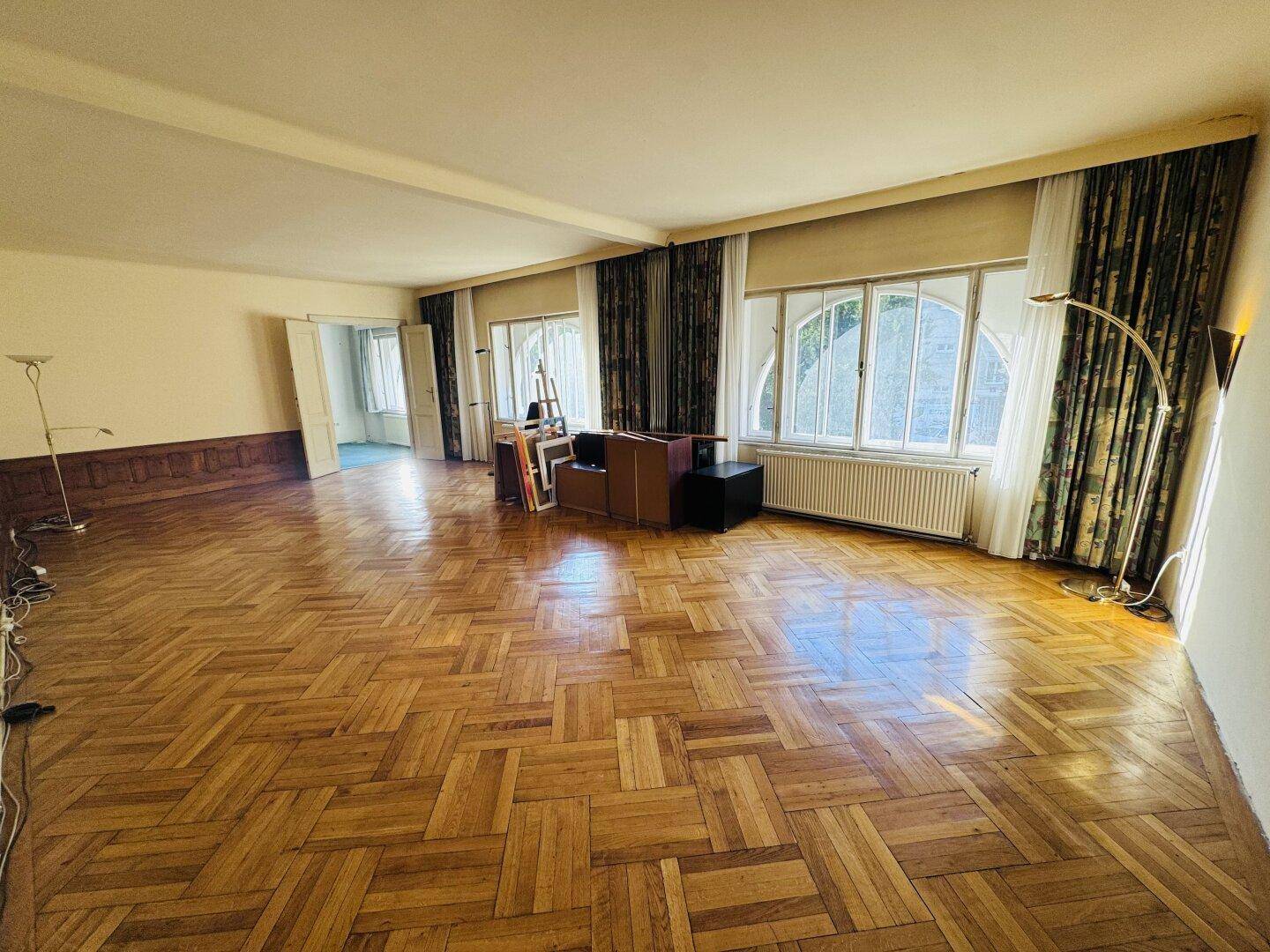 Helles Wohnzimmer mit klassischem Fischgrätparkett und großen Fenstern mit Vorhängen.