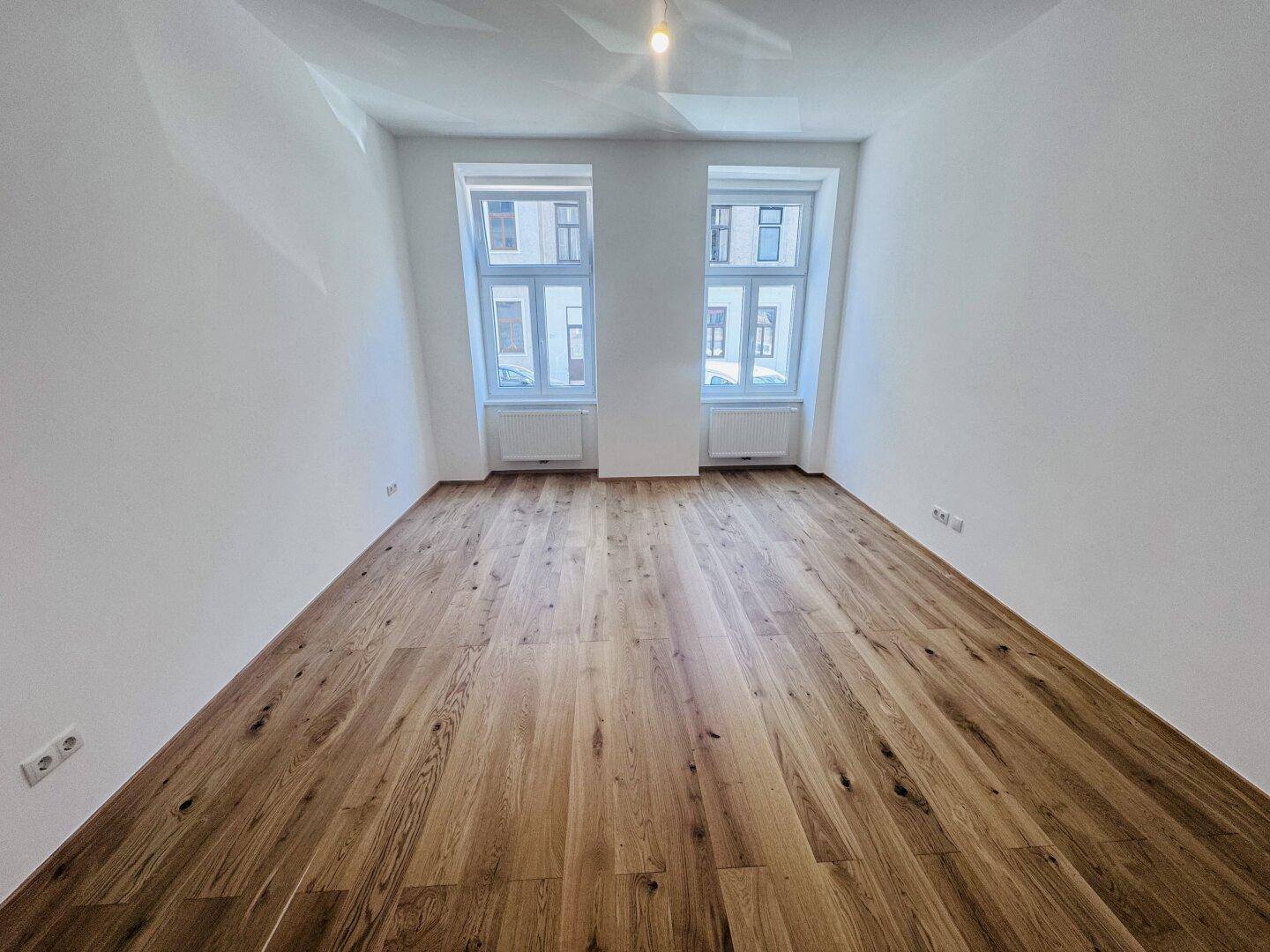 Geräumiges Zimmer mit hellem Holzboden und zwei Fenstern, die den Raum gut belichten.