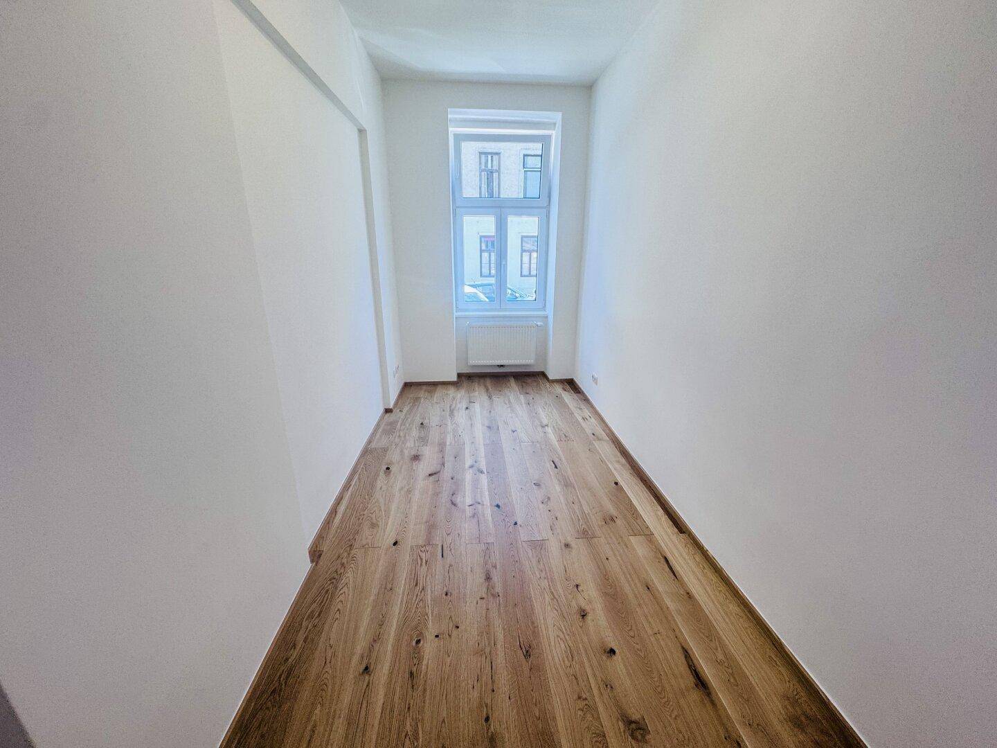 Langes, helles Zimmer mit Holzboden und einem großen Fenster, das den Raum erhellt.