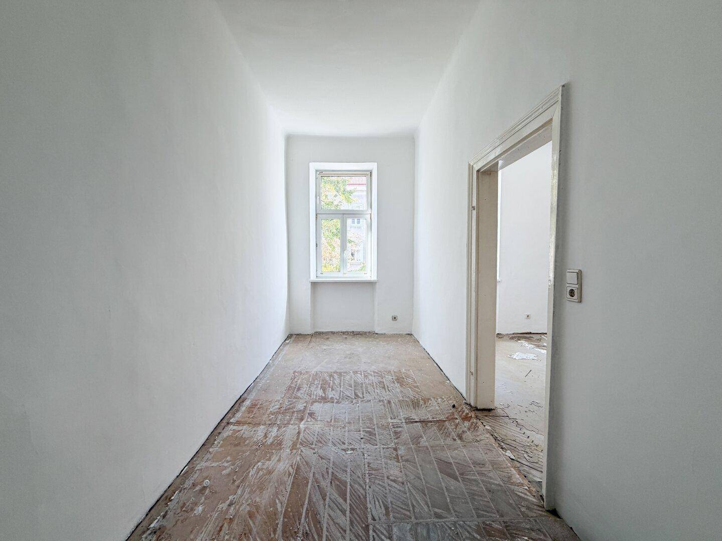 Schmaler Flur mit einem Fenster und alten Holzdielen, der zu weiteren Räumen führt.