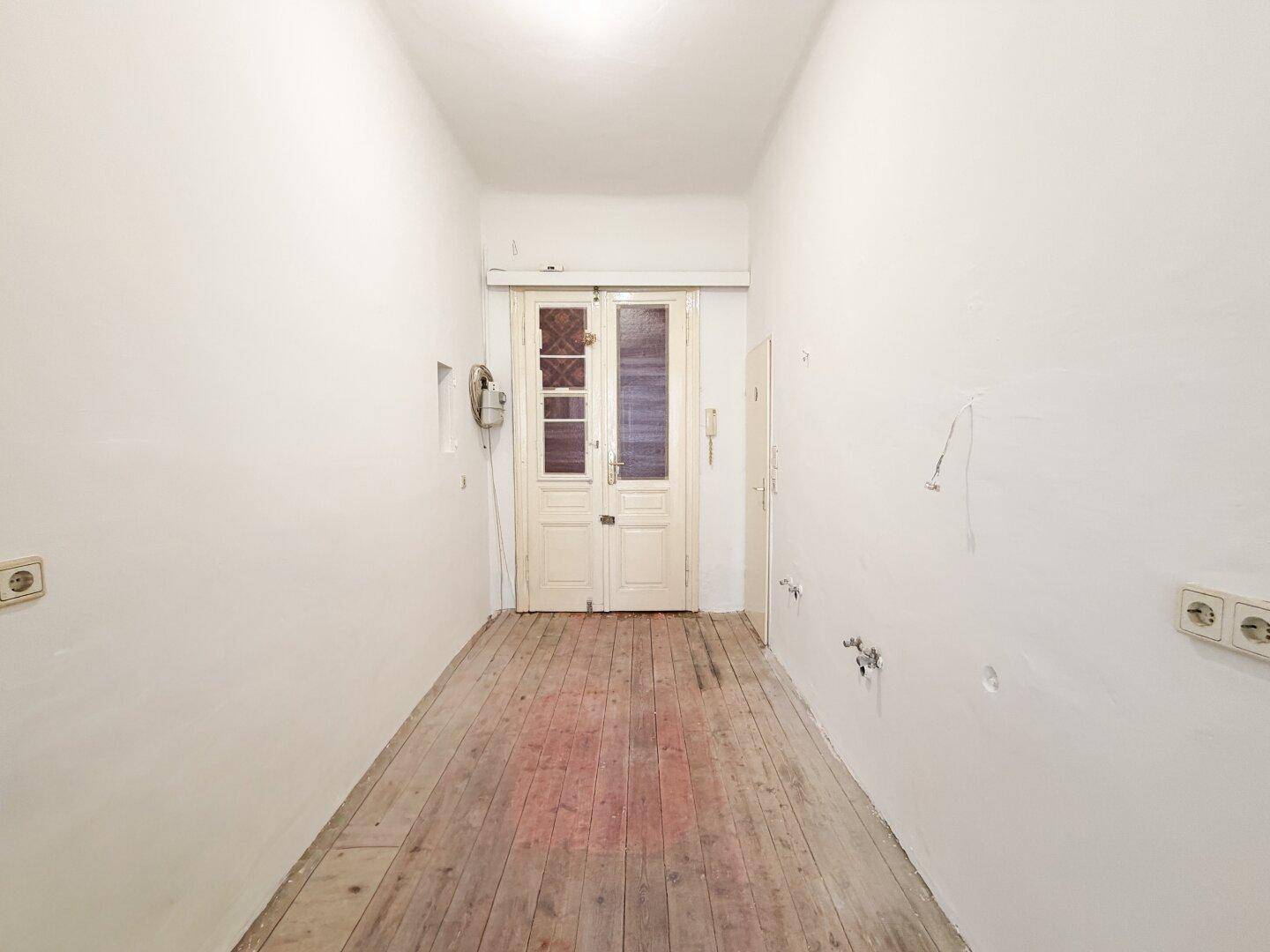 Langer Flur mit Holzdielen und einer Doppeltür am Ende, die Licht hereinlässt.