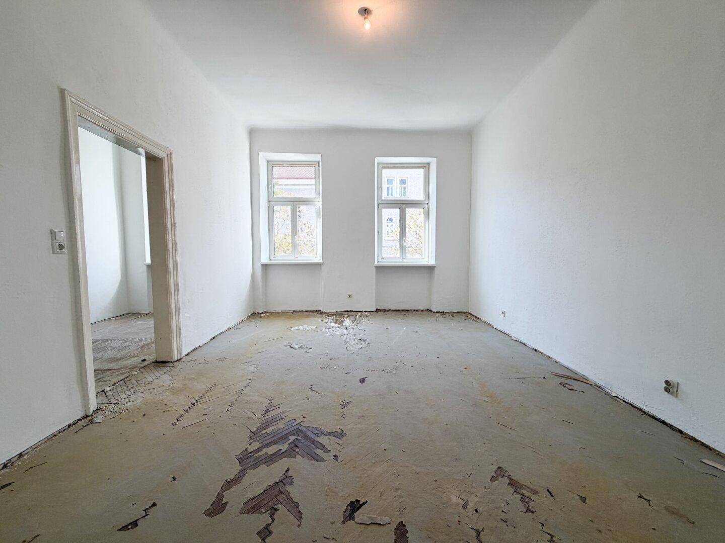 Heller, leerer Raum mit zwei Fenstern und beschädigtem Boden, bereit zur Renovierung.