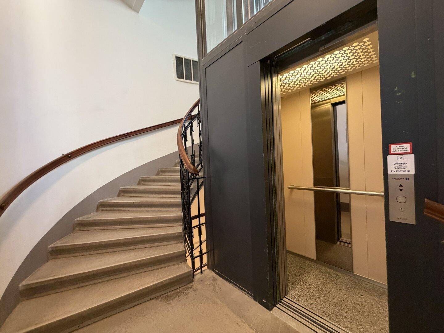 Treppenhaus mit geschwungener Steintreppe und modernem Aufzug.