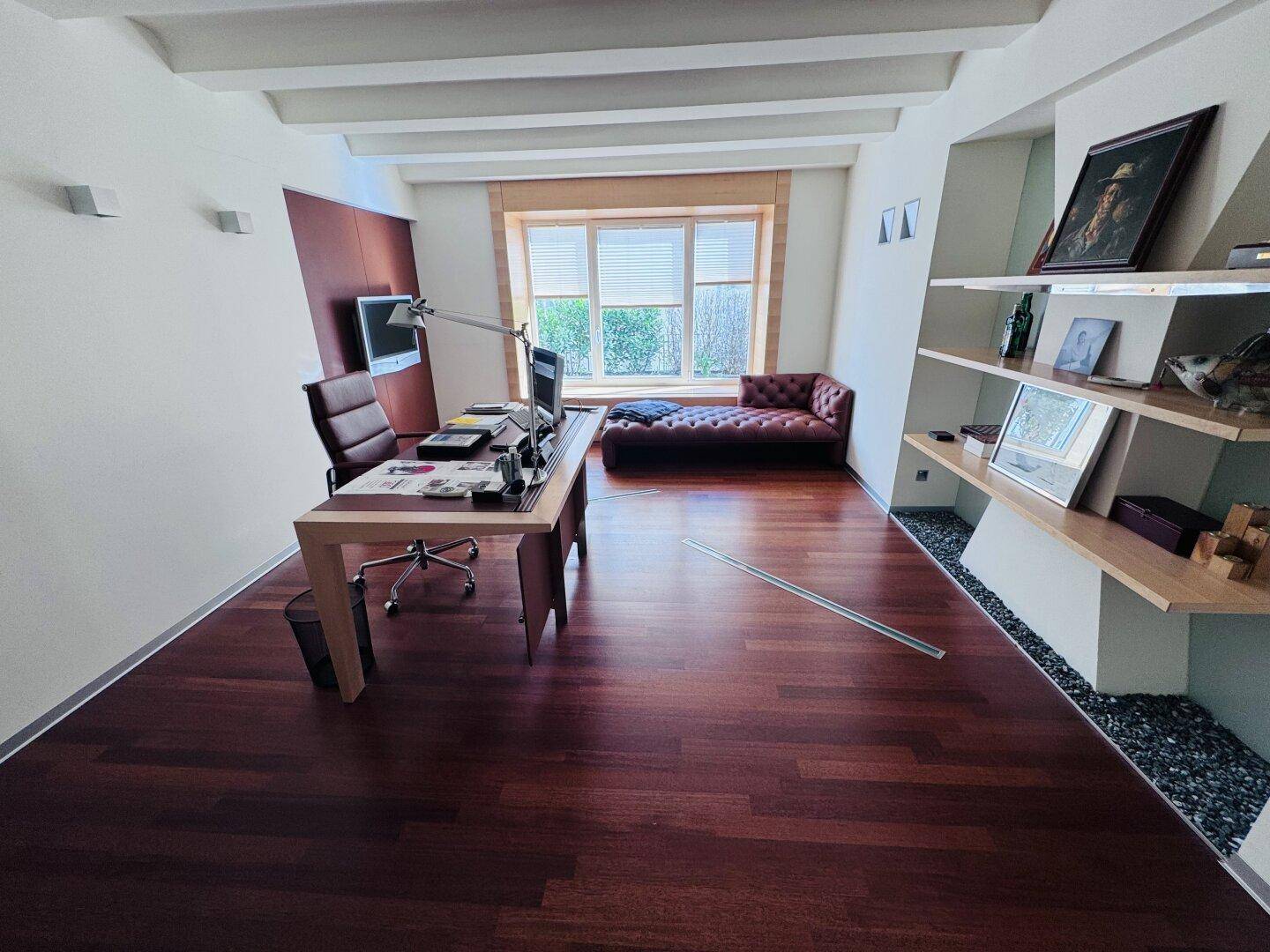 Ein elegantes Büro mit Holzboden, einem großen Schreibtisch und einem bequemen Sofa am Fenster.