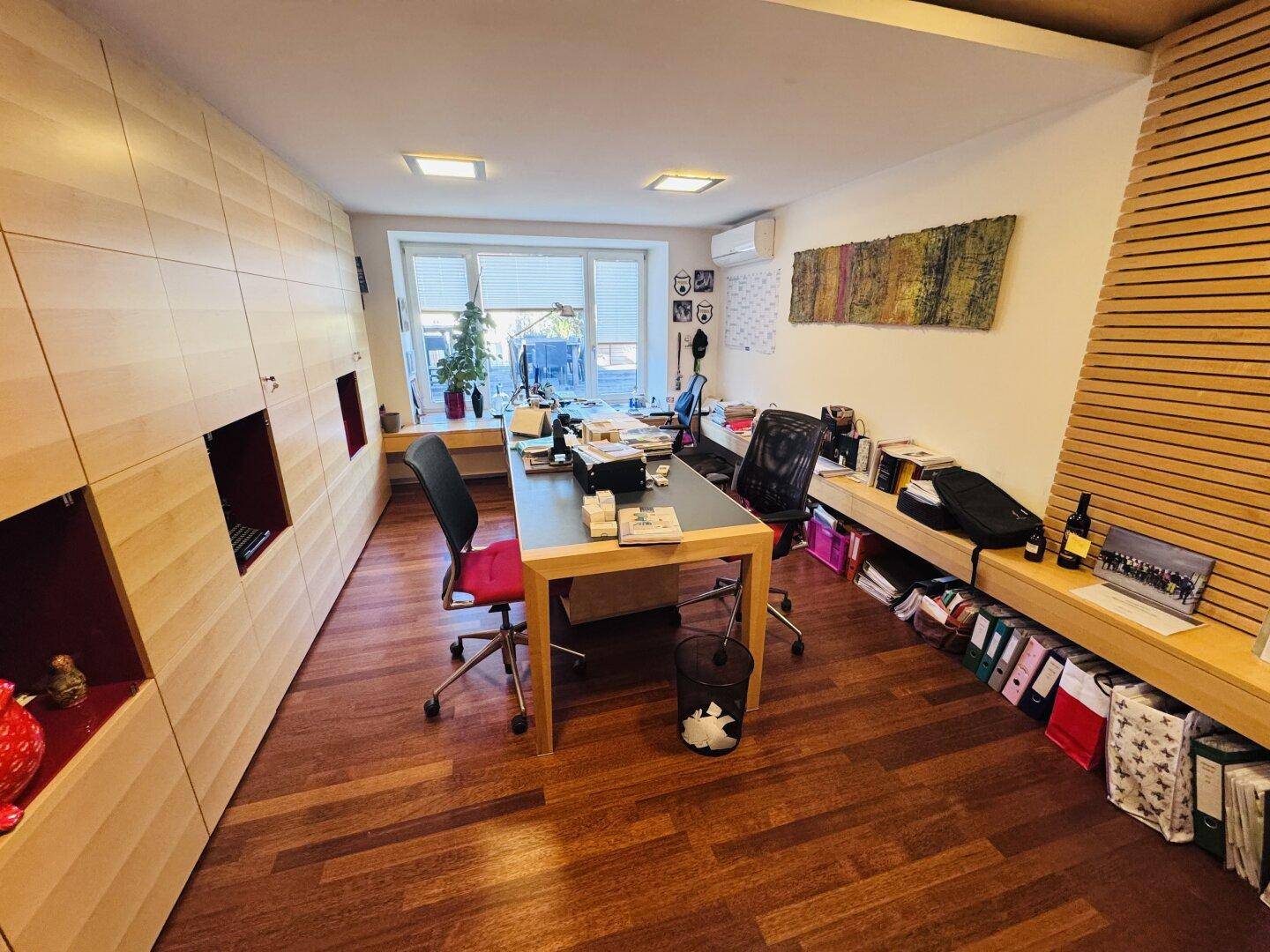 Ein modernes Büro mit Holzboden, einer langen Schrankwand und einem großen Fenster für viel Licht.