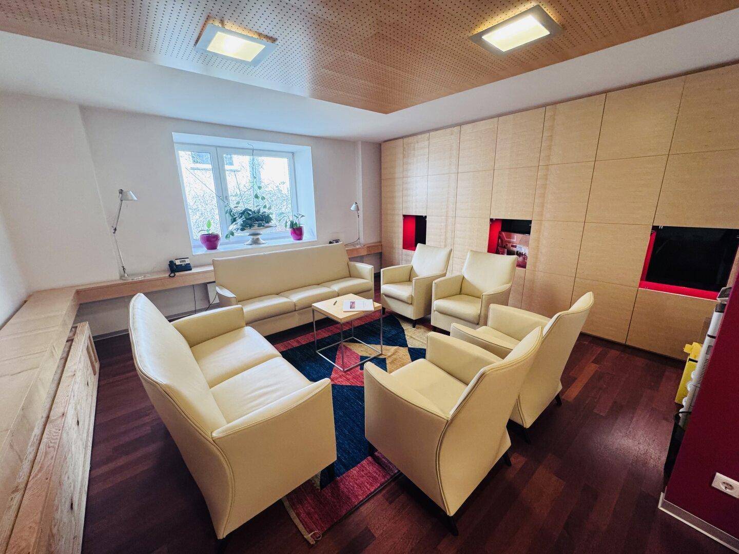 Gemütlicher Wartebereich mit beigen Ledersofas, Sesseln und einem bunten Teppich vor einem Fenster.