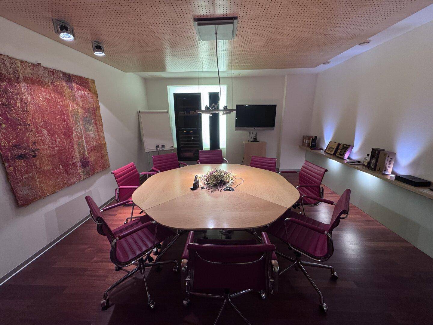 Konferenzraum mit großem achteckigem Holztisch, roten Stühlen und einem Fernseher an der Wand.