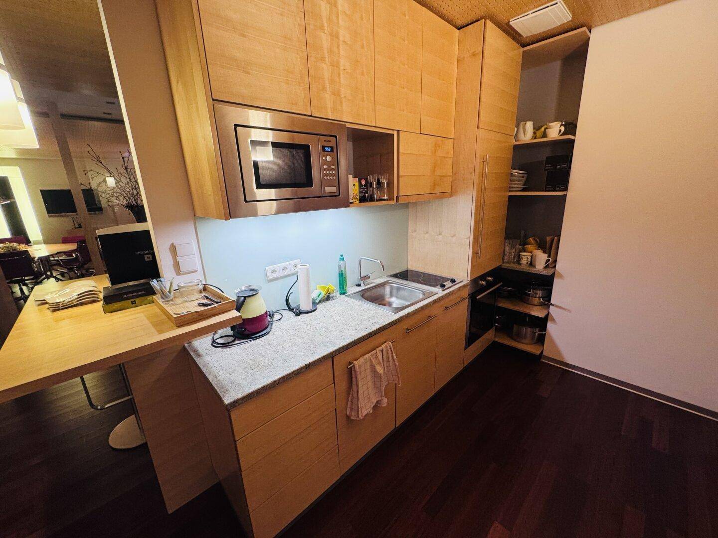 Kleine Küchenzeile mit Holzschränken, Mikrowelle und Spüle, ideal für Büronutzung.