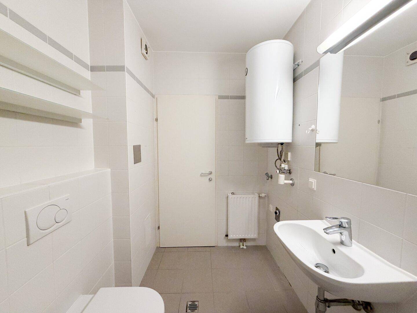 Funktionales Badezimmer mit weißen Fliesen, Waschbecken, WC und Warmwasserboiler.