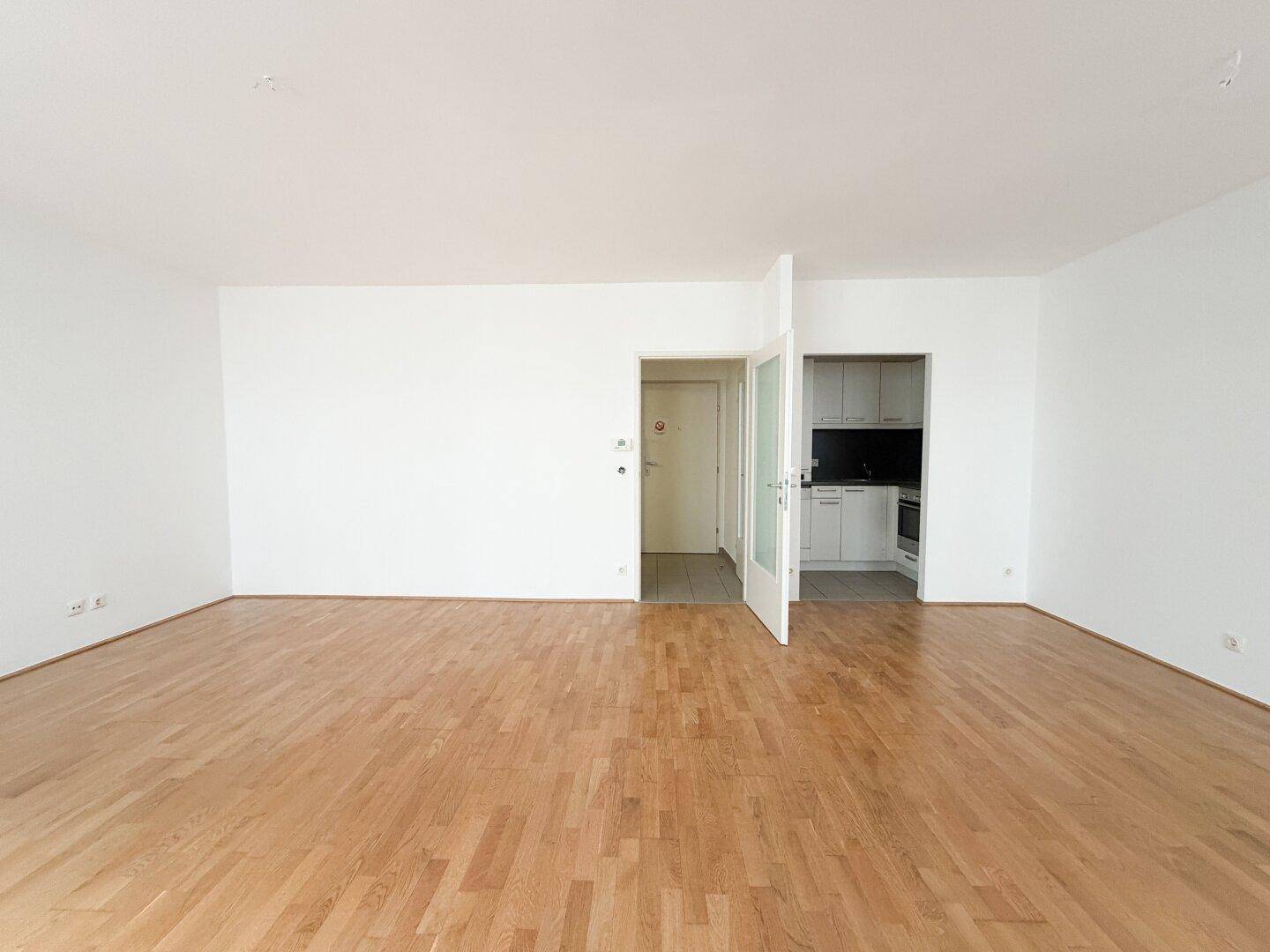 Geräumiges Wohnzimmer mit Parkettboden, weißen Wänden und Zugang zu einer kleinen Küche.