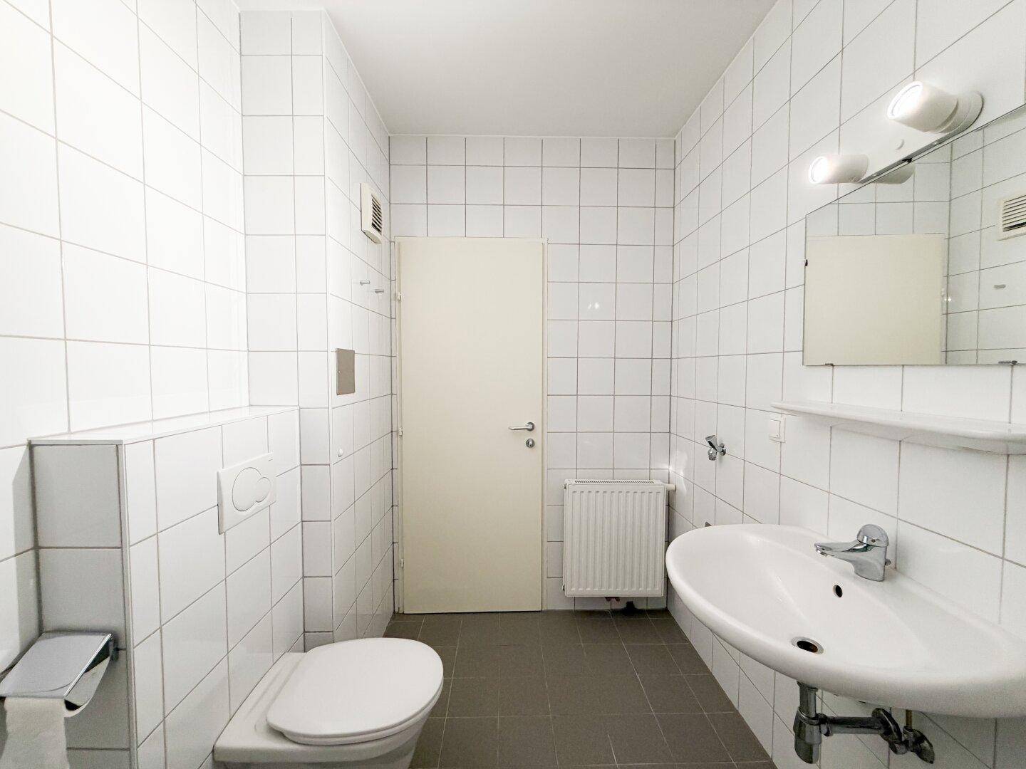 Helles Badezimmer mit WC, Waschbecken und weißen Wandfliesen, ausgestattet mit Heizkörper.