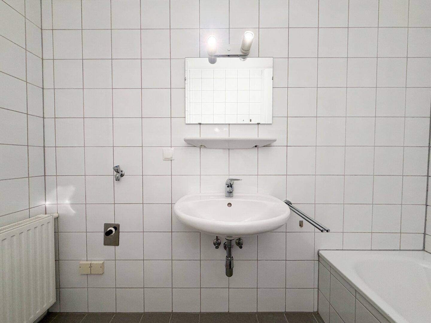 Einfaches Badezimmer mit Waschbecken und Badewanne, komplett gefliest in Weiß, mit Heizkörper.