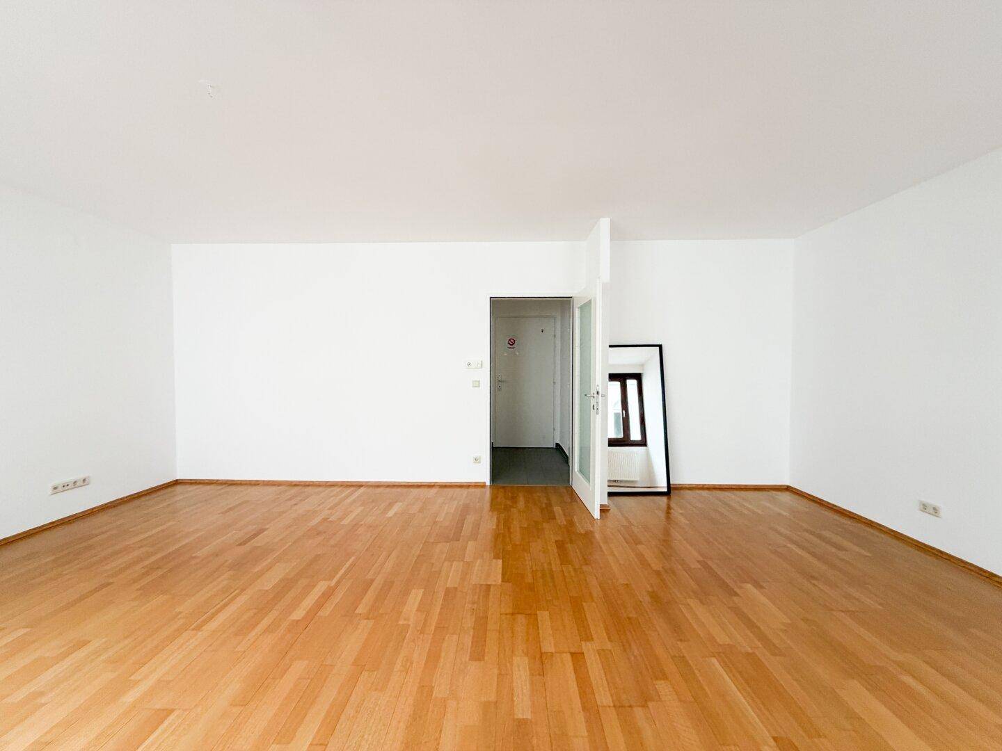 Geräumiges Wohnzimmer mit Parkettboden und weiß gestrichenen Wänden, bietet viel Platz.
