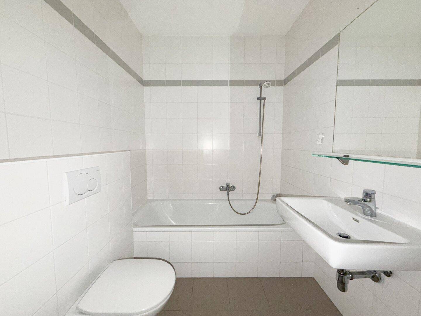 Helles Badezimmer mit weißen Fliesen, Badewanne mit Duschfunktion, WC und Waschbecken.
